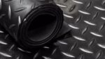 A imagem mostra um rolo de piso de borracha com textura de diamante em fundo preto. O material é resistente e possui um padrão antiderrapante, ideal para áreas comerciais e industriais onde a segurança e a durabilidade são prioridades. O piso está enrolado, evidenciando sua flexibilidade e possibilidade de fácil instalação em diferentes ambientes.