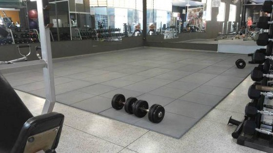 Uma área de ginástica com piso de borracha cinza interligado sobre o piso principal. No centro, um par de halteres repousa sobre o tapete, indicando uma zona de treinamento de força. Ao redor da área, há equipamentos de ginástica, pesos livres e paredes espelhadas que refletem o espaço e a luz natural que entra pelas janelas.