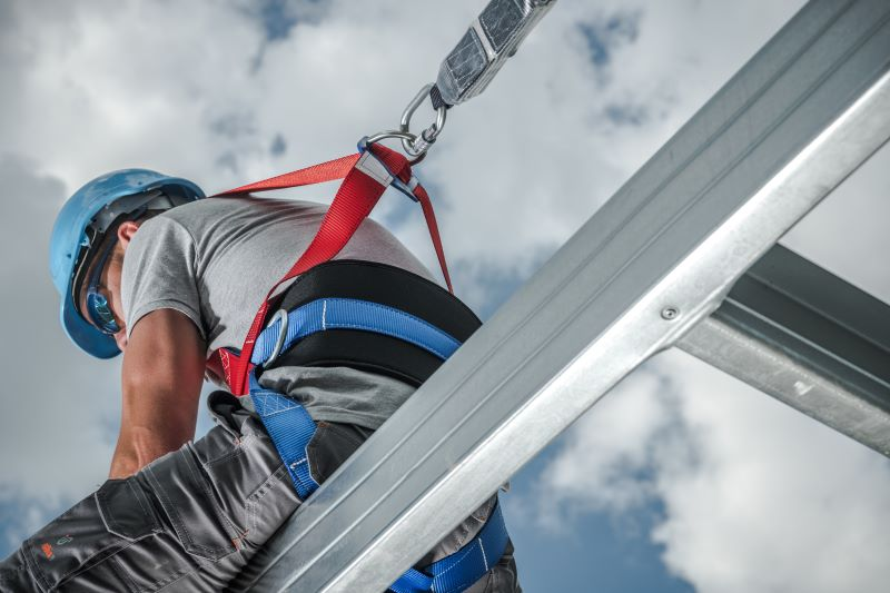 Um trabalhador da construção civil usando um capacete de segurança azul e um cinto de proteção contra quedas de corpo inteiro está preso a uma estrutura metálica com um cordão de segurança.