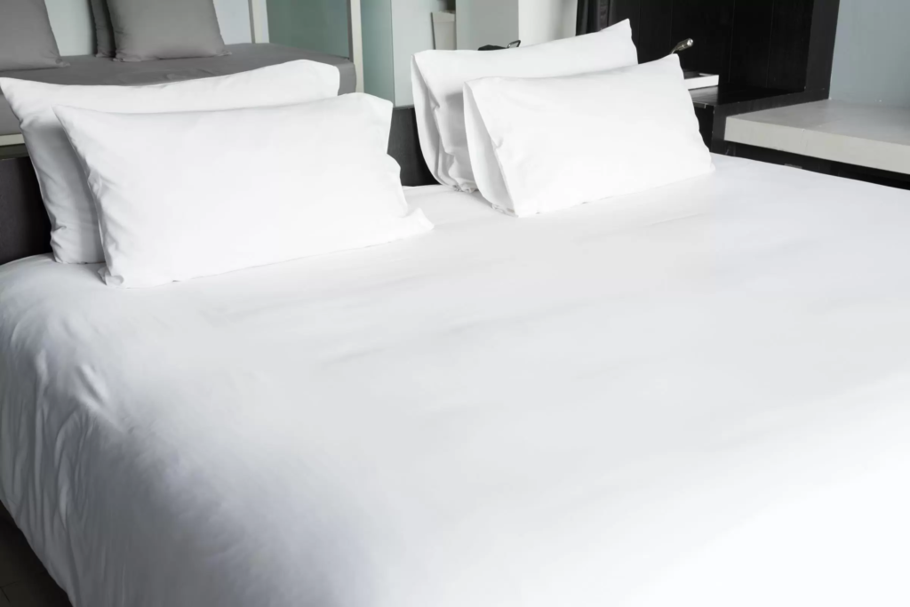 Uma cama forrada com lençol branco e com quatro travesseiros de poliuterano com fronhas brancas.