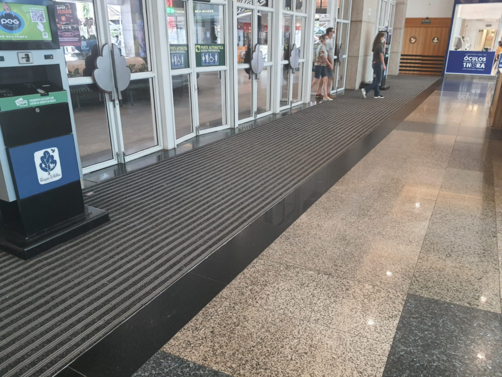 Visão de um corredor em um espaço comercial, com portas de vidro ao fundo. No chão, há um tapete de entrada com faixa antiderrapante, projetado para reter sujeira e umidade, ajudando a manter a área limpa e segura.
