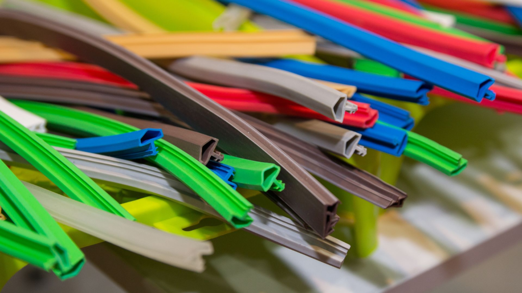 Uma composição de tiras de borracha e plástico em várias cores, como verde, azul, marrom, vermelho e amarelo, dispostas de forma desordenada. As tiras possuem diferentes formatos e texturas, com alguns perfis sendo mais altos e outros mais planos. A imagem evidencia a diversidade de usos desses materiais, que podem ser aplicados em vedação ou acabamento em diferentes produtos industriais.