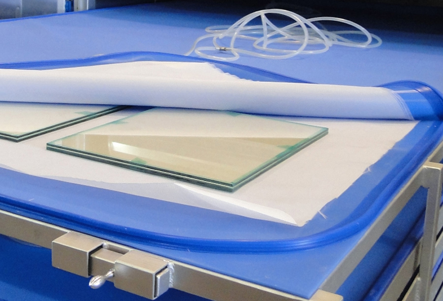 Superfície de trabalho de laminação a vácuo com folhas de vidro dispostas. Uma bolsa de silicone está parcialmente desenrolada sobre os vidros, pronta para a laminação. Tubos transparentes estão visíveis, indicando o sistema de vácuo. O ambiente é organizado, com uma mesa de trabalho de fundo azul.
