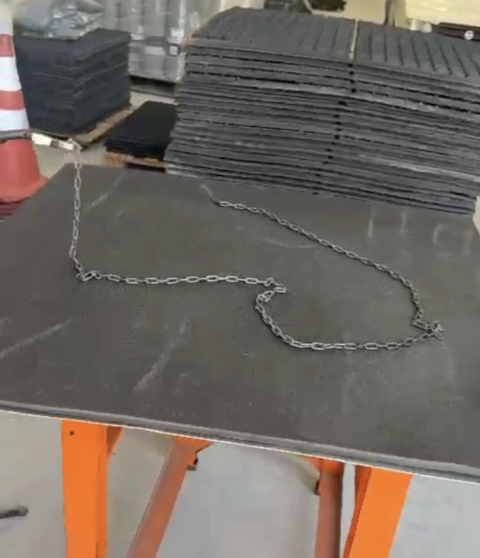 Imagem de uma mesa de trabalho com um estrado emborrachado escuro, sobre o qual está disposta uma corrente de metal. Ao fundo, empilhados, estão diversos estrados isolantes, enquanto um cone de sinalização está parcialmente visível à esquerda.