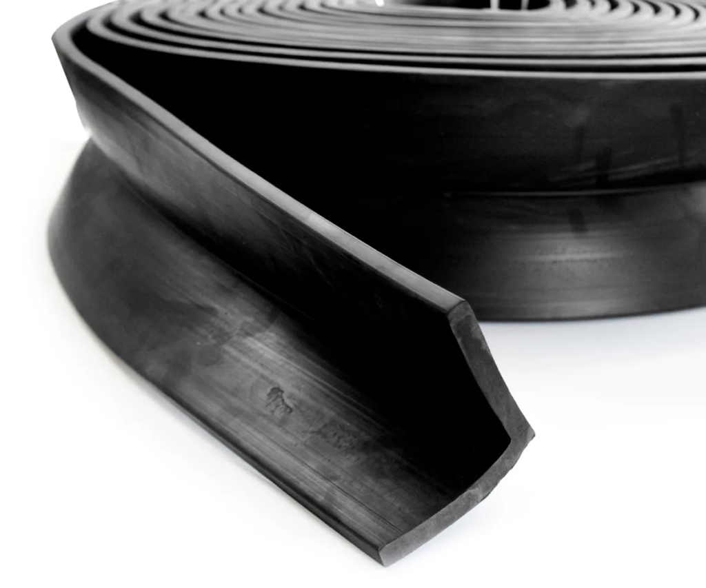 Imagem de uma cantoneira de borracha preta, em formato de L, com uma das extremidades cortada em ângulo, mostrando sua textura lisa e resistente. A peça é envolvida por várias bobinas adicionais ao fundo, também de borracha ou material similar, sugerindo que faz parte de um conjunto maior ou de um estoque de materiais de proteção e acabamento para edificações ou industrial.