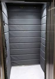 Interior de um elevador de carga acolchoado com painéis de parede acolchoados em cinza, projetados para proteger tanto a carga quanto o elevador durante o transporte. O acolchoamento cobre todas as três paredes internas, e o piso é uma superfície de cor clara, contrastando com os tons mais escuros do acolchoamento. A imagem foi tirada do d