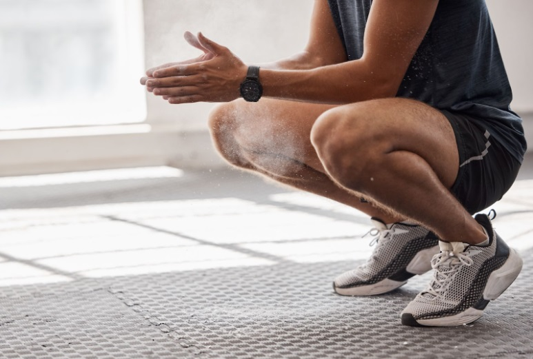 Uma pessoa vestindo roupas esportivas e tênis agachada no piso texturizado de uma academia de EVA, preparando-se para um treino esfregando giz nas mãos. A luz natural forte de uma janela próxima destaca o padrão do piso e cria uma atmosfera motivacional de treinamento.