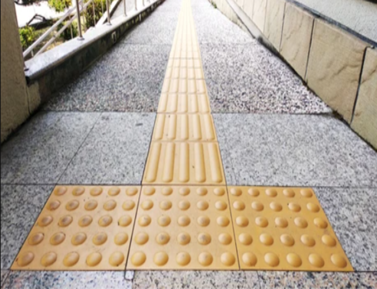 Imagem de uma calçada com piso tátil em amarelo. O piso possui diferentes texturas: várias placas retangulares com ranhuras em uma faixa central e placas quadradas com pontos em toda a superfície. Essas diferentes texturas indicam que a calçada é adaptada para auxiliar quem tem deficiência visual na orientação e navegação pelo espaço. A calçada é construída com materiais de concreto, cercada por uma parede de pedra na direita e uma cerca de proteção na esquerda.