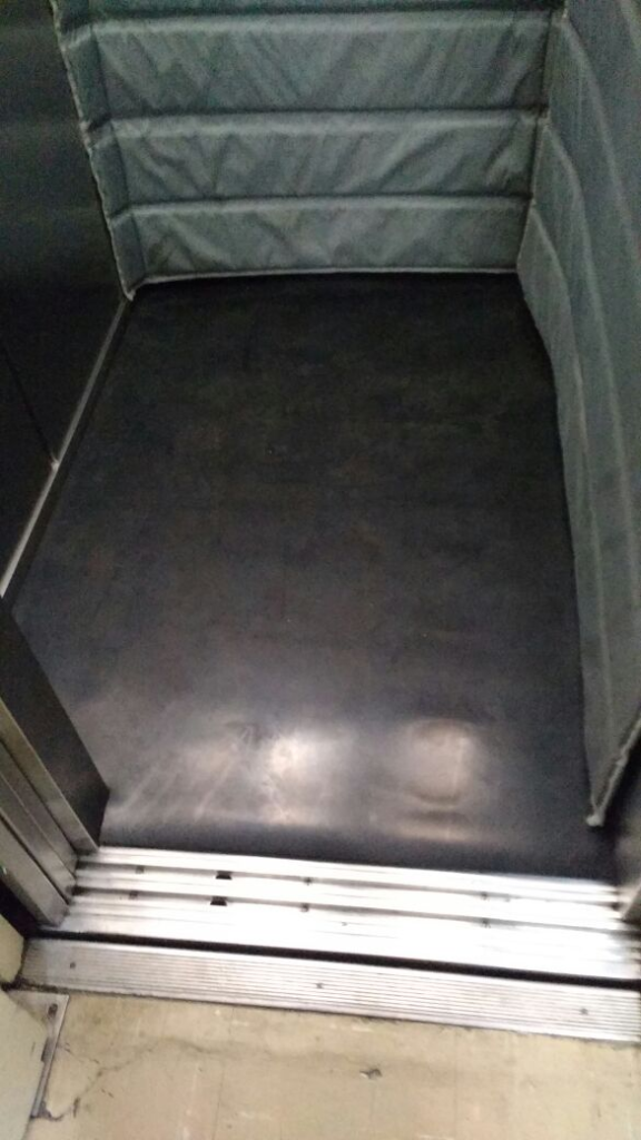 Imagem de um interior de elevador, com piso de aparência escura e lisa, possivelmente de borracha ou outro material resistente. A porta do elevador está parcialmente aberta, revelando o piso externo, que é de cor clara e possui uma linha de trilho metálico na soleira. Na parte de trás do elevador, há uma parede com painel acolchoado acolchoado de tom cinza, com três faixas horizontais, provavelmente para proteção ou acabamento. A iluminação reflete suavemente sobre o piso, indicando uma superfície limpa e bem conservada.