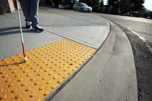 Uma pessoa com bengala acompanha o deslocamento ao lado de uma faixa de piso tátil de alerta amarela instalada na calçada de um bairro, próximo ao cruzamento de uma rua. A faixa de piso tátil tem textura de pontos elevados em relevo, acionando alertas sensoriais para pessoas com deficiência visual, sinalizando a aproximação de uma área de risco ou de mudança de direção na passagem.