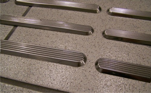 "Imagem que mostra várias peças de piso tátil de cor prata, com linhas elevadas e texturizadas, dispostas sobre uma superfície de cor cinza, geralmente usadas para sinalização de orientação para pessoas com deficiência visual."