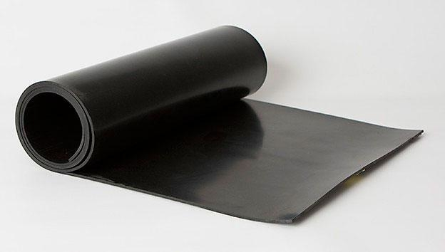 Imagem de um rolo de material preto, parcialmente desenrolado, que parece ser uma folha de borracha ou similar. O material tem uma textura lisa e é utilizado geralmente para fins de vedação, isolamento ou proteção em aplicações industriais e domésticas.