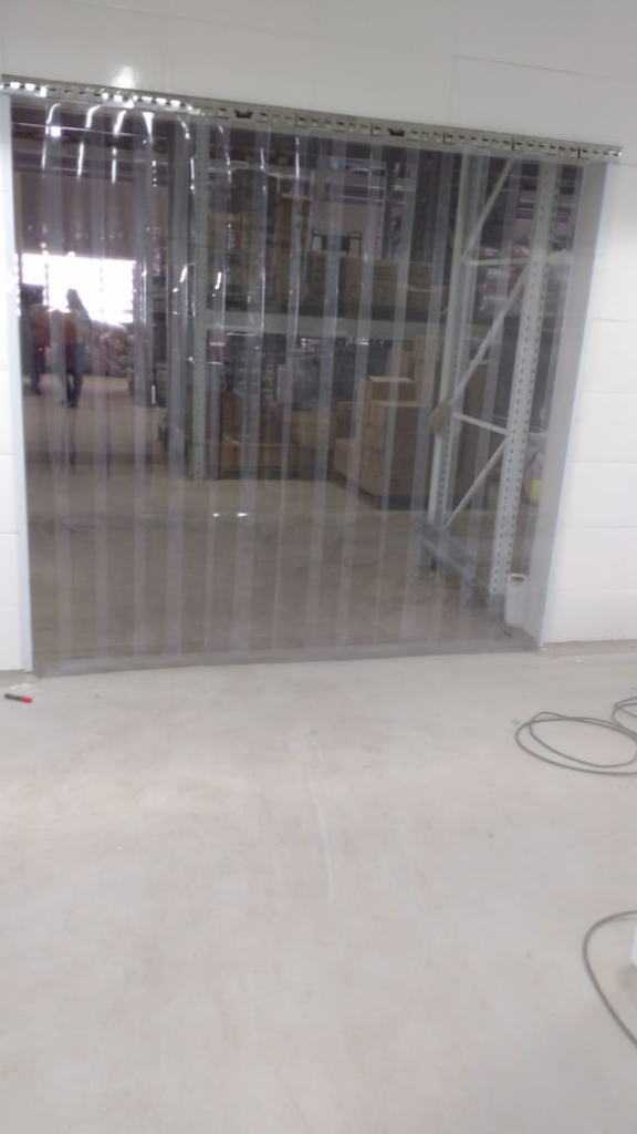 Na imagem, é possível ver uma cortina de PVC transparente instalada em um galpão. Ela separa o ambiente interno do estoque, ajudando a manter a limpeza e a temperatura, além de permitir a passagem de pessoas e empilhadeiras sem abrir portas.