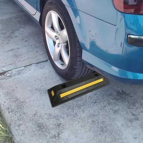 Um carro azul está estacionado com o pneu traseiro apoiado em um batente de borracha preta. O batente possui uma faixa amarela refletiva para visibilidade e está posicionado sobre uma superfície de concreto, servindo como uma barreira para impedir que o veículo se mova ainda mais para trás.
