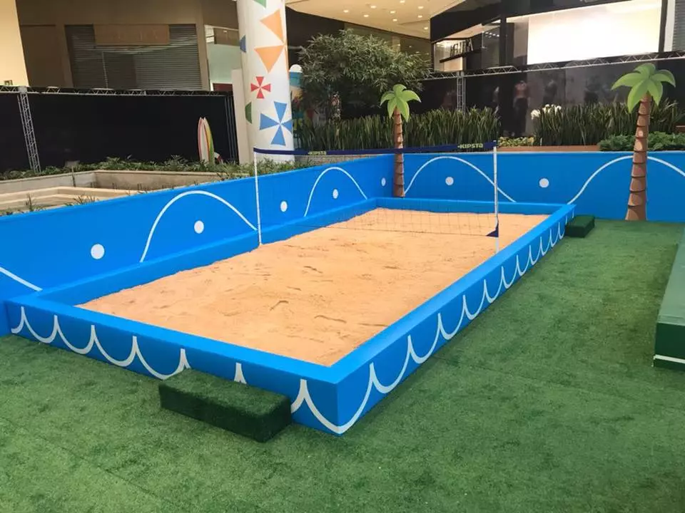 Uma quadra de areia em tamanho pequeno, rodeada por uma estrutura azul decorada com ondas brancas, simulando o tema do mar. A quadra tem linhas brancas marcando o campo de jogo e duas redes pequenas, indicando que também pode ser usada para jogos de vôlei ou futebol de areia. O local está cercado por grama artificial e há árvores decorativas ao fundo, criando um ambiente divertido e temático.
