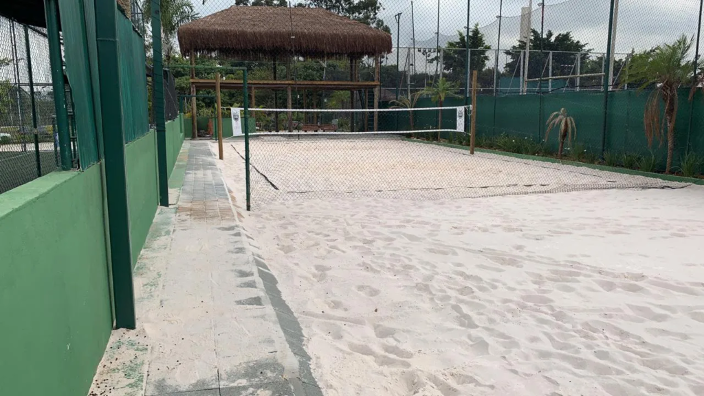 Uma quadra de areia de esporte, delimitada por uma cerca e paredes verdes, com sinalização para jogo, possivelmente de vôlei ou areia. A área possui uma faixa de passeio ao lado, feita de concreto, e no fundo, há uma área sombreada com uma cabana de thatch e árvores ao redor, criando um ambiente ao ar livre confortável para práticas esportivas.
