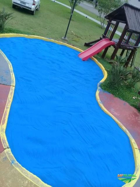 Um balanço de areia em formato de lago, com a superfície de areia azul, rodeado por uma faixa amarela. Ao fundo, há uma casinha de brinquedo com escorregador rosa e uma área com grama e árvores, criando um ambiente de lazer infantil ao ar livre.