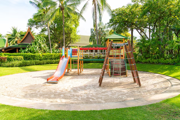 Uma estrutura de playground com escorregador em um parque ao ar livre. O entorno é cheio de árvores, arbustos e grama bem cuidado, criando um ambiente natural e convidativo para as crianças brincarem. A área de recreação é cercada por uma superfície de areia, proporcionando segurança durante as atividades.