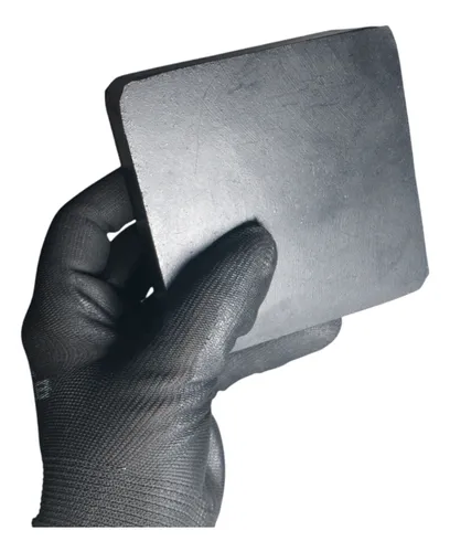 Uma mão usando luva preta segura um bloco retangular de borracha, com uma borda arredondada e superfície lisa. O objeto está próximo à câmera, em posição vertical, com o canto superior inclinado. A textura da luva contrasta com o acabamento suave da peça, que parece pesado e sólido.