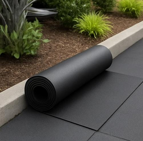 A imagem mostra um rolo de material de borracha preto, provavelmente um tipo de piso ou isolamento, colocado ao lado de uma linha de piso pavimentado de cor escura. O ambiente parece ser externo, com algumas plantas e vegetação ao fundo. O material de borracha provavelmente é utilizado para revestimento, isolamento ou proteção de superfícies contra impacto, vibração ou intempéries, sendo uma opção prática e durável para áreas externas ou de convivência ao ar livre.