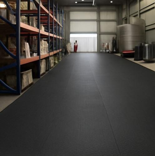 A imagem mostra o interior de um galpão ou depósito equipado com uma viarly de piso de borracha preta estendida ao longo do espaço. A área é bem organizada, com prateleiras carregadas de caixas e produtos ao lado esquerdo e grandes tanques ou reservatórios ao fundo à direita. O piso de borracha proporciona uma superfície sólida, resistente e segura, ideal para ambientes industriais ou de armazenamento, oferecendo proteção contra impactos e maior segurança para trabalhadores e equipamentos.