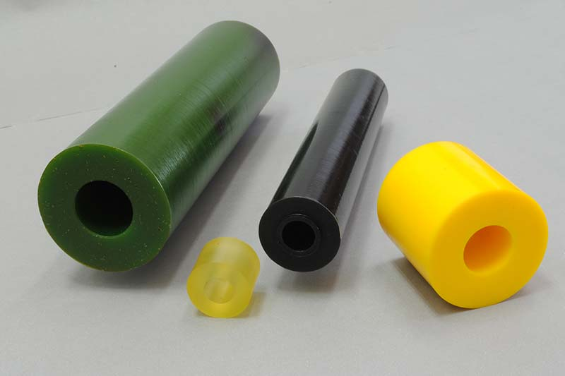 Quatro barras cilíndricas de borracha com furos centrais, de diferentes tamanhos e cores — verde, preto, amarelo translúcido e amarelo opaco  colocadas sobre uma superfície cinza claro, apresentando variações de material e diâmetro.