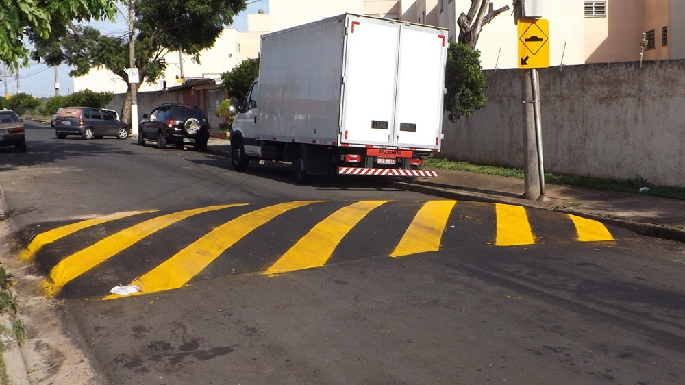 Rua com lombada estreita pintada em faixas diagonais amarelas e pretas. Um caminhão branco está estacionado ao lado da calçada, parcialmente encostado na faixa de pedestres pintada. No fundo há carros estacionados, uma árvore, muro baixo e sinal de trânsito amarelo com mudança de direção. A cena sugere obra ou ajuste viário com sinalização de alerta.