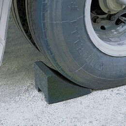  uma roda de caminhão apoiada sobre um bloco triangular preto colocado entre a roda e o solo. A superfície ao redor é de cascalho ou brita fina em tom claro, sugerindo área de manobra ou reparo viário. A imagem enfatiza a posição de apoio da roda, possivelmente durante manutenção ou controle de peso.