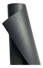 Rolos de material em cor cinza-escuro, com textura lisa e brilho suave. Um rolo está parcialmente desenrolado, revelando superfícies planas em camadas; o conjunto parece ser de uma folha ou lâmina de material sintético espesso, dobrando-se levemente nas bordas. A iluminação suave realça as tonalidades acinzentadas e a geometria angular das folhas.