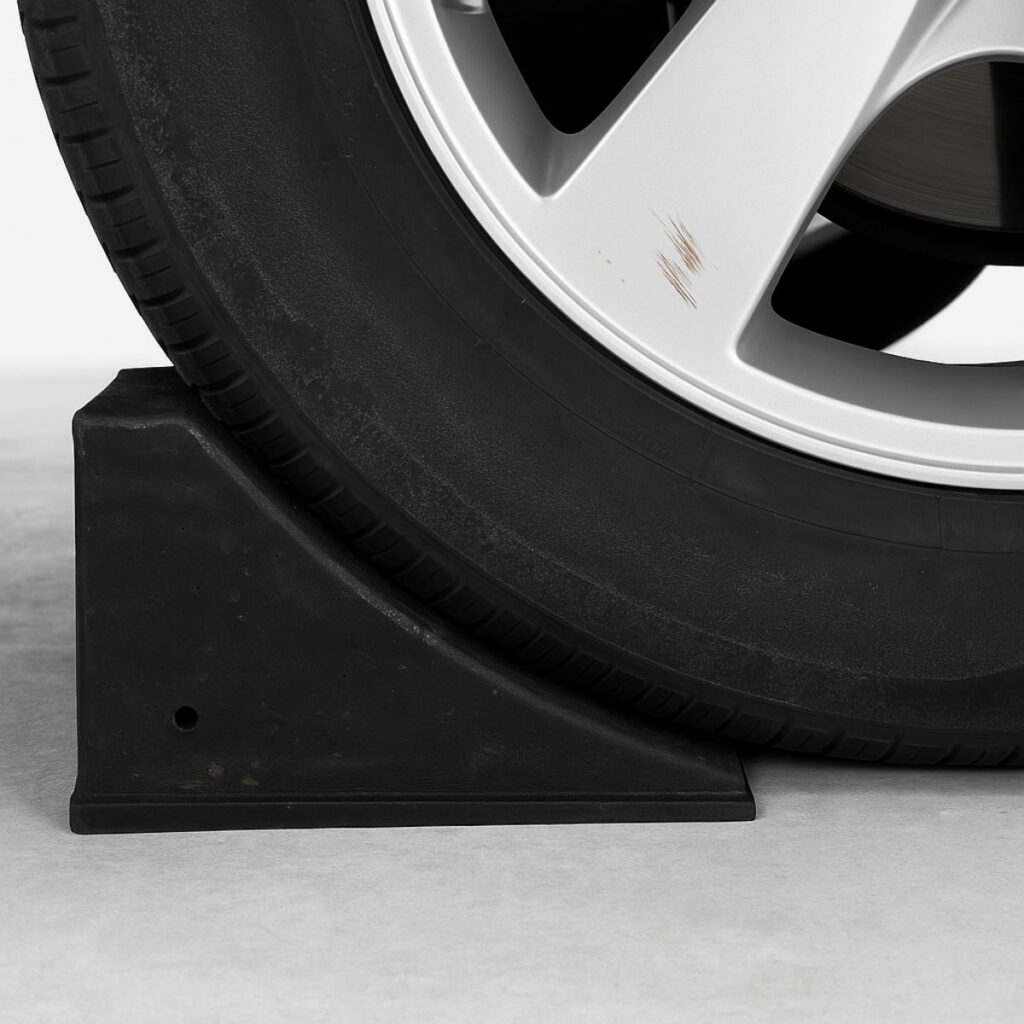 Rodas de veículo apoiadas sobre uma rampa ou calço de borracha preta, com a face lateral do aro prata visível. A superfície está com marcas de desgaste e o piso ao fundo é claro e liso. A composição sugere uma cena de proteção ou suporte para reparo de pneu, destacando a aderência entre o pneu e o suporte.