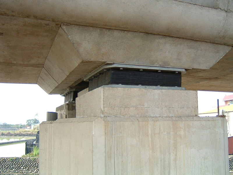 Pilar de concreto sob uma ponte, com capas e blocos empilhados formando a base de sustentação. A estrutura mostra cabeçotes de apoio de borracha ou material elastomérico entre a ponte e o pilar, possivelmente para absorção de vibrações. Cenário externo com água ao fundo e vegetação, iluminação natural.