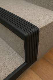 Detalhe de uma escada com uma peça de proteção preta de borracha que envolve a borda e cria várias ranhuras paralelas. A peça está fixada na borda de uma escada em pedra clara, com piso de madeira visível na parte inferior. A cor predominante é o preto da peça de vedação, contrastando com o tom bege da pedra e o piso de madeira.