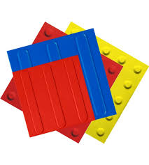 Quatro peças de piso tátil coloridas sobrepostas: uma placa vermelha com ranhuras no centro, uma placa azul com saliência vertical, uma placa amarela com pinos, tudo disposto em ângulo sobre uma superfície branca. As cores vibrantes e o relevo característico das peças criam um arranjo lúdico e geométrico.