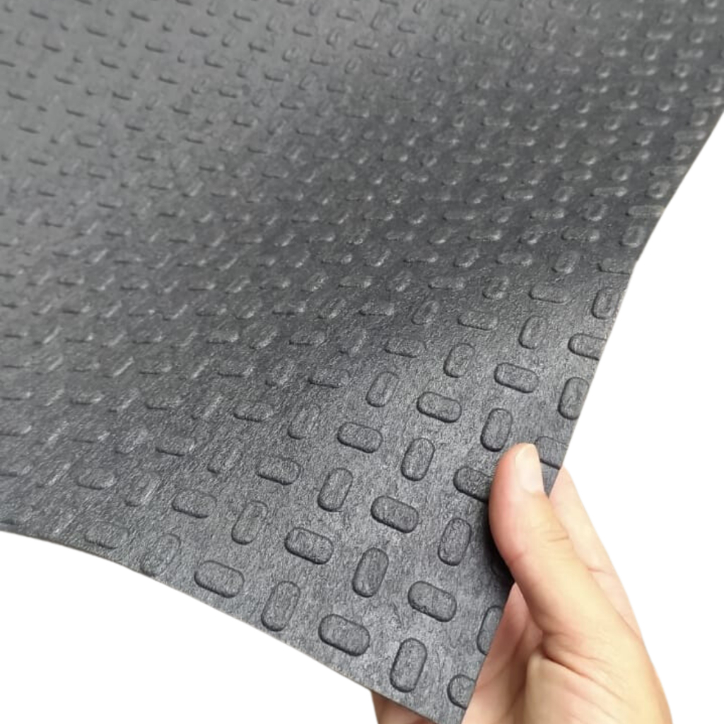 Imagem de uma mão segurando uma folha ou pedaço de material preto com padrão texturizado em relevo, composto por pequenas elipses alongadas repetidas. A superfície é lisa e impermeável, com acabamento acetinado. A mão está pegando a borda direita, revelando o contorno curvado da peça.