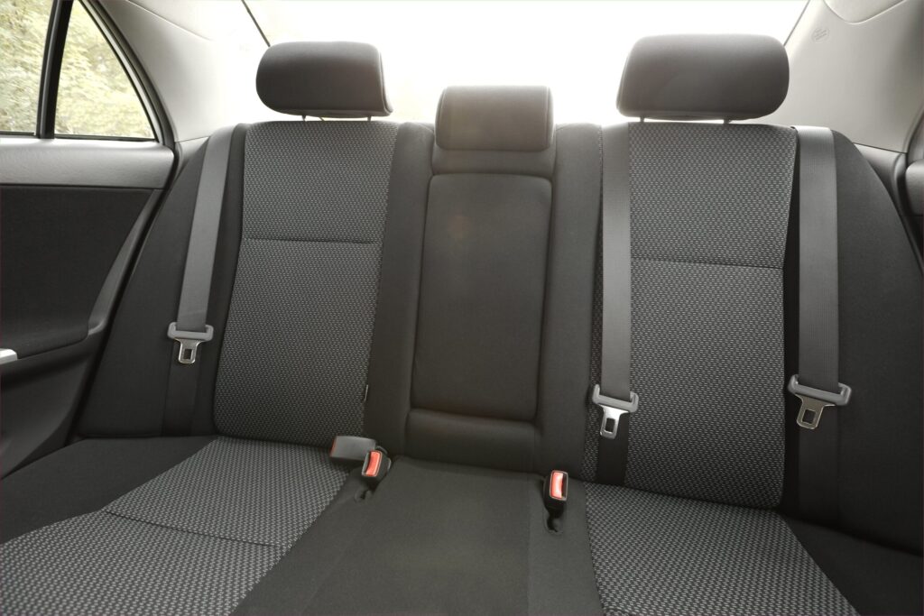Interior de carro visto de trás, banco traseiro com encosto central recuado, três cintos de segurança pretos e fivelas prateadas. Estofamento cinza-escuro com padrão de malha, material acolchoado e costuras, bordas pretas, com apoios de cabeça ajustáveis ao fundo. Lanternas de luz natural entram pela janela lateral.