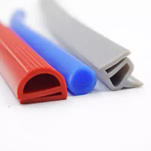 A imagem apresenta três perfis de vedação de silicone coloridos e distintos, dispostos diagonalmente sobre um fundo branco. Da esquerda para a direita, o primeiro é um perfil vermelho em formato de "D", com estrias longitudinais na parte superior. O segundo é um perfil azul redondo e liso. O terceiro é um perfil cinza claro com um formato de "U" ou "J", com uma aba interna, mostrando um design mais complexo.