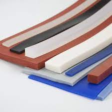 A imagem exibe uma variedade de perfis e folhas de silicone em diferentes cores e espessuras, dispostos em camadas e em ângulos sobre um fundo claro. Predominam tons de vermelho-tijolo, azul, cinza, branco e preto. As peças incluem tiras retangulares finas, barras mais espessas e folhas planas, todas com superfícies lisas, destacando a diversidade de formatos disponíveis para vedação e outras aplicações industriais.