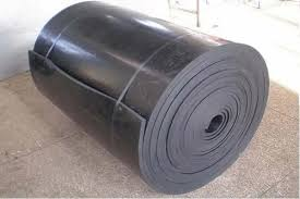 A imagem exibe um grande rolo de material de borracha preta, possivelmente butílica, com várias camadas visíveis em sua lateral. O rolo está posicionado na diagonal sobre um piso que parece ser de concreto ou cimento em um ambiente industrial ou de armazenamento. A superfície da borracha é lisa, e algumas tiras escuras, possivelmente fitas de amarração, envolvem o rolo para mantê-lo compacto.