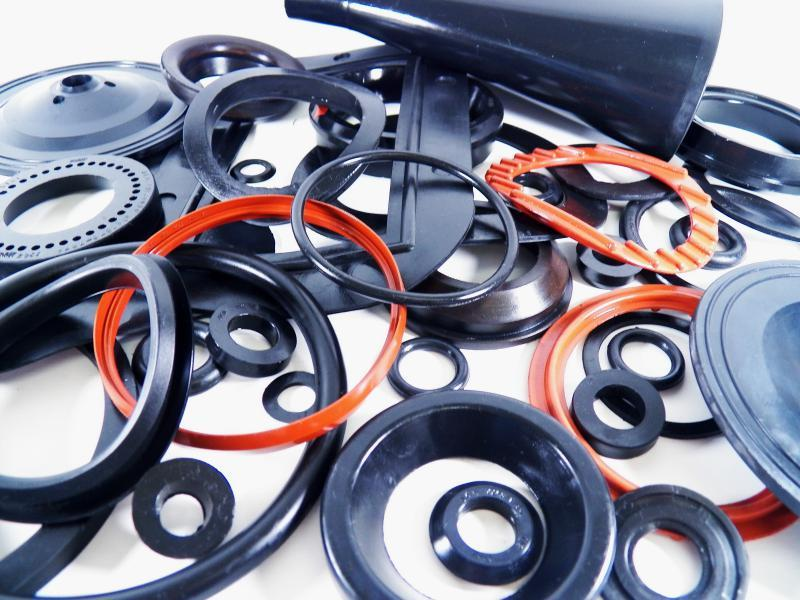 Conjunto de anéis de vedação, vedantes e componentes de borracha e plástico espalhados desordenadamente sobre uma superfície clara. Os anéis variam em diâmetro, espessura e cor, com tons pretos e vermelhos, alguns com furos centrais visíveis. Peças côncavas, cônicas e roldanas podem ser vistas ao fundo, sugerindo um ambiente de oficina ou linha de montagem. A cena transmite a ideia de peças de reposição, manutenção de equipamentos e montagem de sistemas hidráulicos ou pneumáticos.
