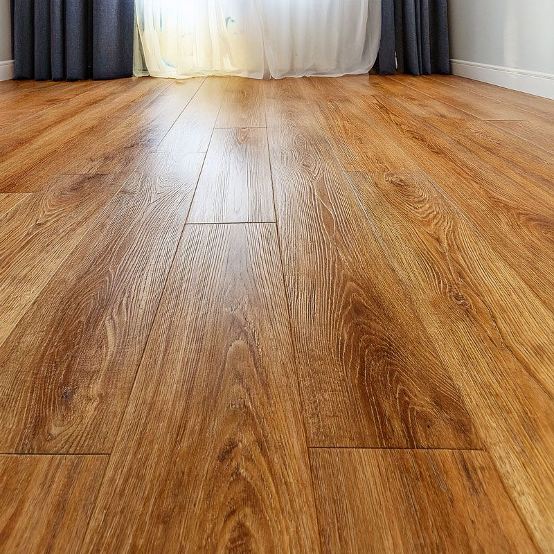 Visão em perspectiva de um piso vinílico instalado em uma sala iluminada. O revestimento possui réguas longas com tonalidade mel e veios de madeira marcados, estendendo-se em direção a uma janela com cortinas cinzas, transmitindo uma sensação de aconchegante e amplitude.