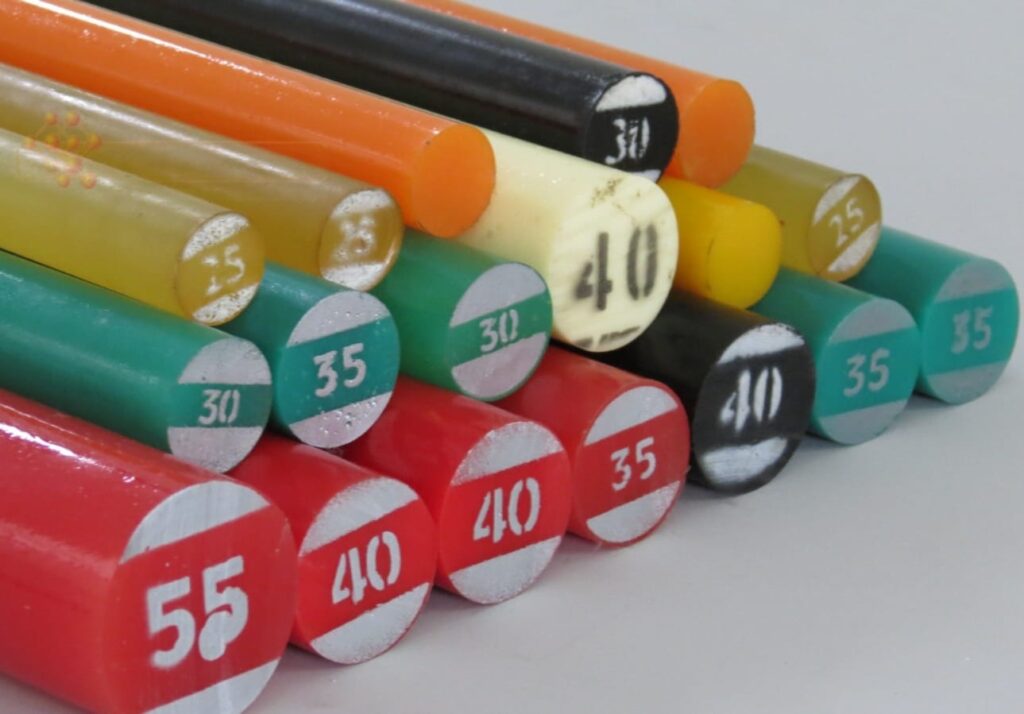 A imagem apresenta uma pilha organizada de bastões cilíndricos industriais, conhecidos tecnicamente como tarugos, feitos de materiais poliméricos como poliuretano (PU), nylon ou plásticos de engenharia similares.