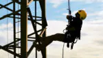 Um trabalhador está descendo de um mastro de torre, suspenso por um sistema de cordas e equipamentos de segurança. Ele usa um capacete amarelo e está vestido com roupas adequadas para trabalho em altura.