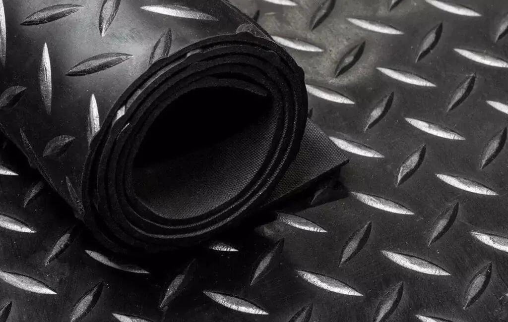 A imagem mostra um rolo de piso de borracha com textura de diamante em fundo preto. O material é resistente e possui um padrão antiderrapante, ideal para áreas comerciais e industriais onde a segurança e a durabilidade são prioridades. O piso está enrolado, evidenciando sua flexibilidade e possibilidade de fácil instalação em diferentes ambientes.