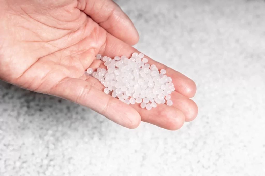 Mão segurando grânulos plásticos transparentes ou levemente esbranquiçados, usados como matéria-prima na produção de plásticos. O fundo está coberto com mais grânulos semelhantes.