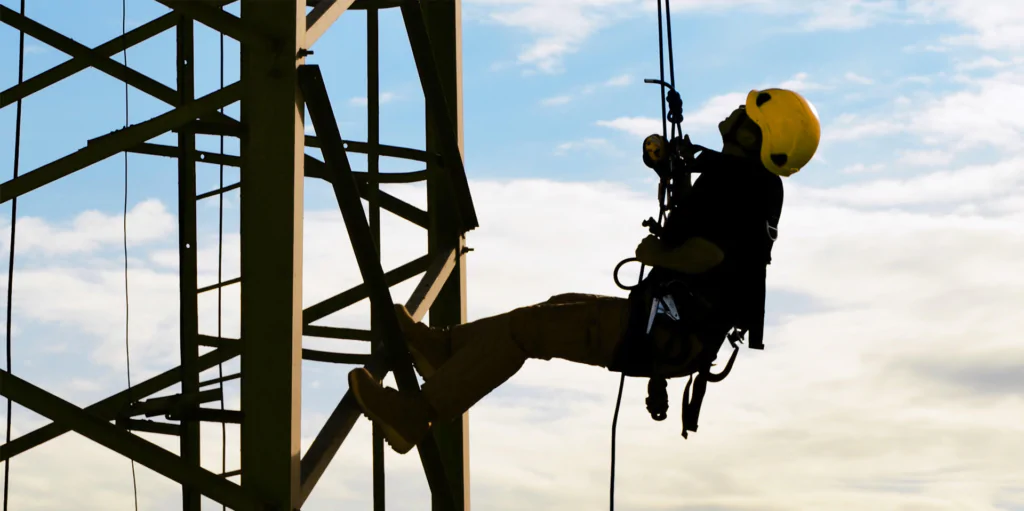 Um trabalhador usando equipamento de segurança, incluindo capacete amarelo e cinto de segurança, está suspenso por cordas enquanto realiza manutenção ou inspeção em uma estrutura metálica alta. 