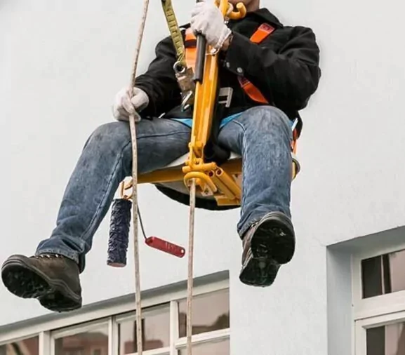 Um trabalhador suspenso por um sistema de acesso por corda está realizando manutenção ou pintura na parede externa de um prédio. 