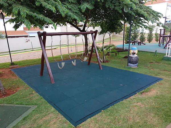 Uma área de recreação infantil com um conjunto de balanços de madeira e três balanços de plástico pendurados em cordas. Os balanços são instalados sobre uma superfície de segurança feita de placas de borracha verde-escuro interligadas. 