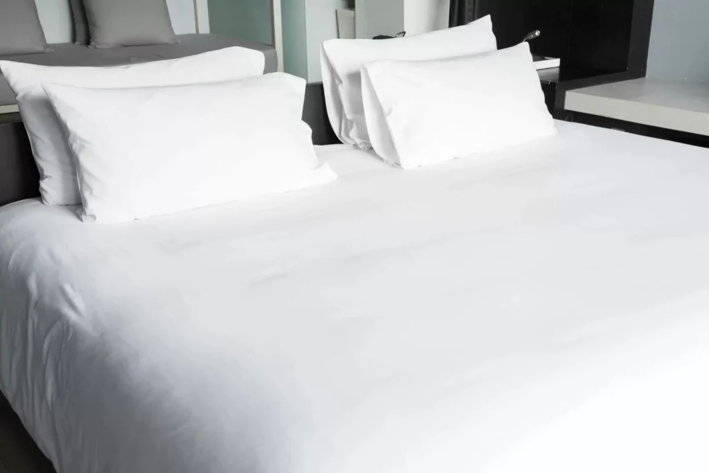 Uma cama forrada com lençol branco e com quatro travesseiros de poliuterano com fronhas brancas.