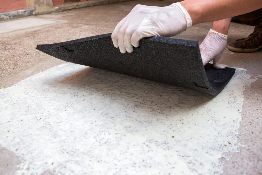 Um trabalhador está colocando uma peça de material espesso e preto sobre uma superfície coberta de uma cola ou adesivo branco. Ele usa luvas brancas e está concentrado na tarefa. 