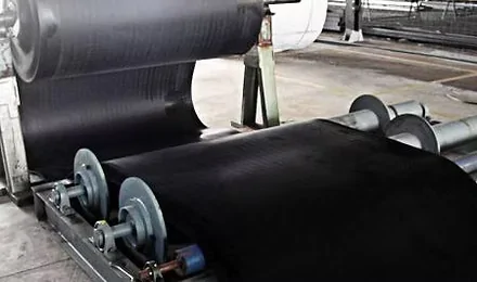 Uma grande máquina industrial processando uma folha contínua de material de borracha preta, com rolos guiando o material pela linha de produção dentro de uma fábrica.